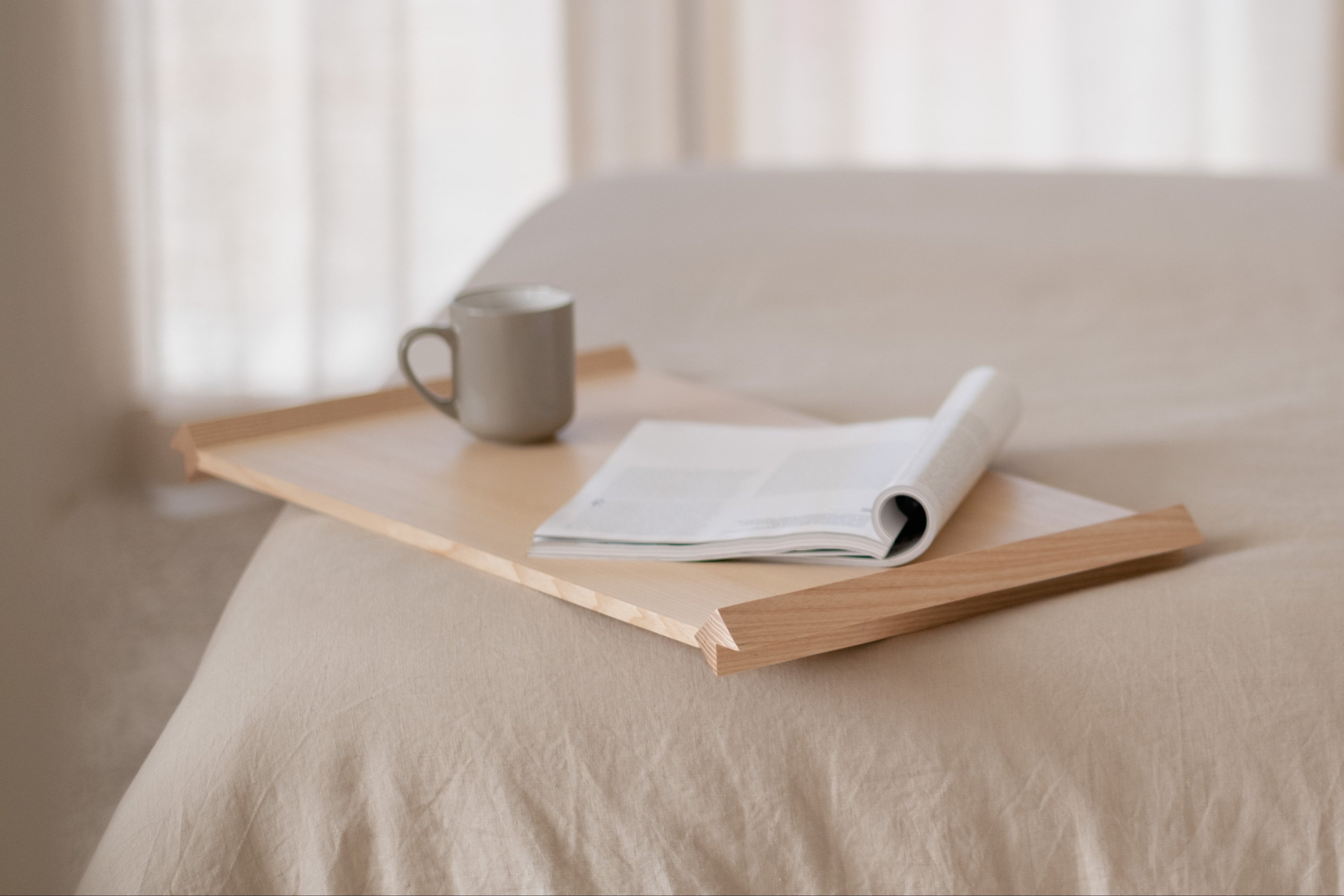 Wooden tray with a mug and book on a bed in a softly lit room