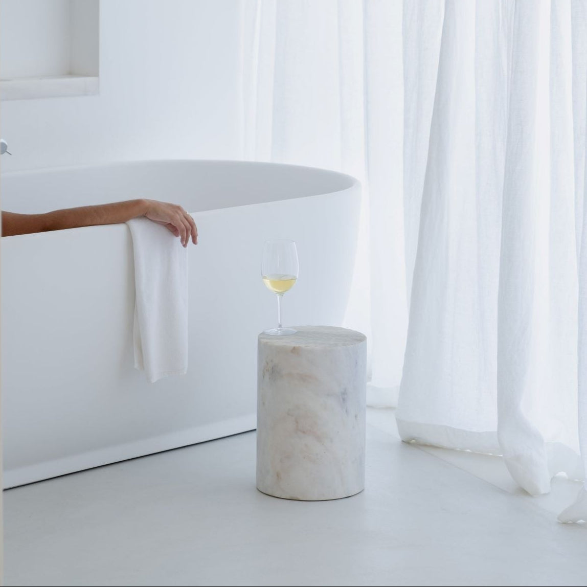 A white minimalist modern side table with a stone texture, named PEDRA, in a room with a white bathtub and curtains in the background.