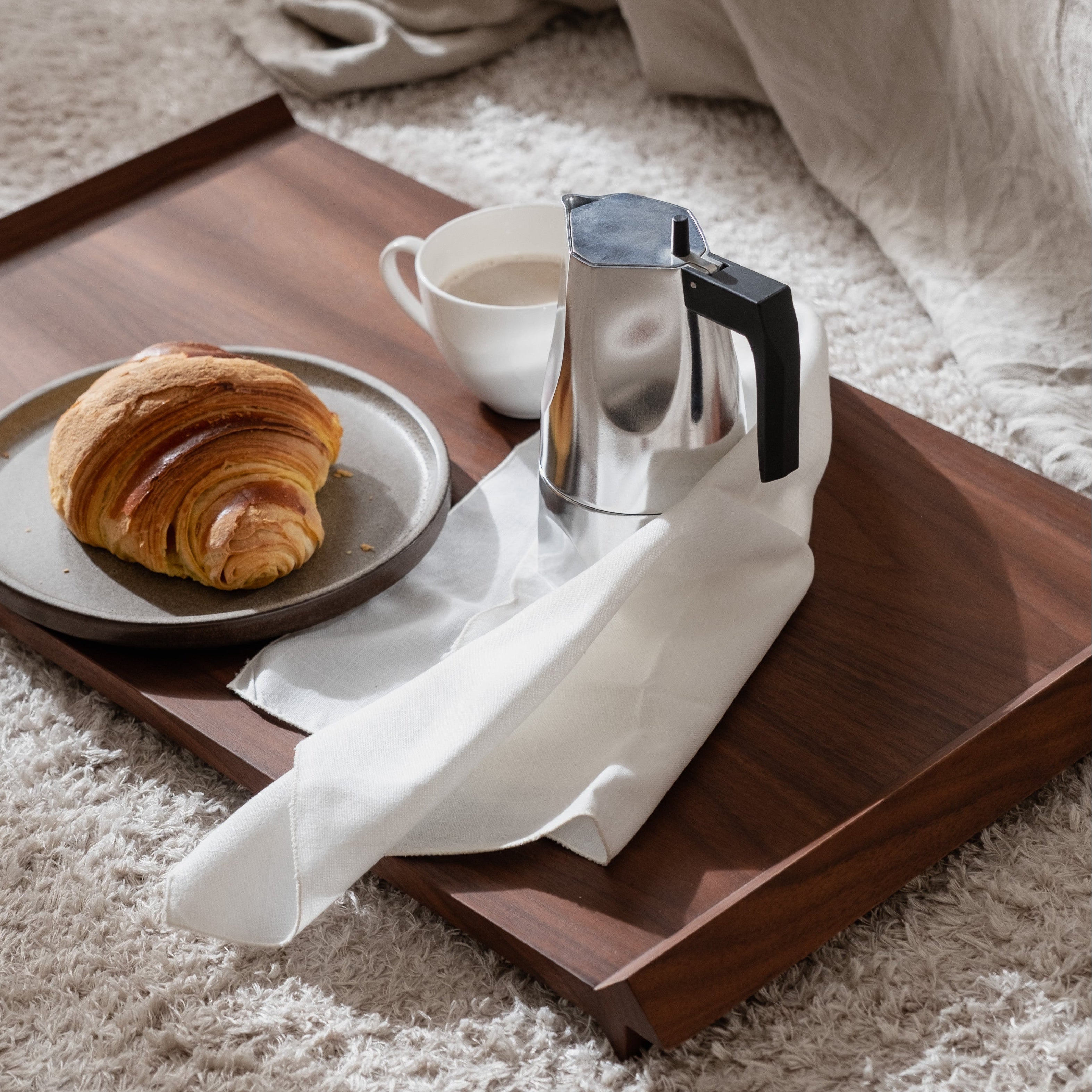Wooden tray with a croissant, coffee, and a pitcher on a carpeted floor.
